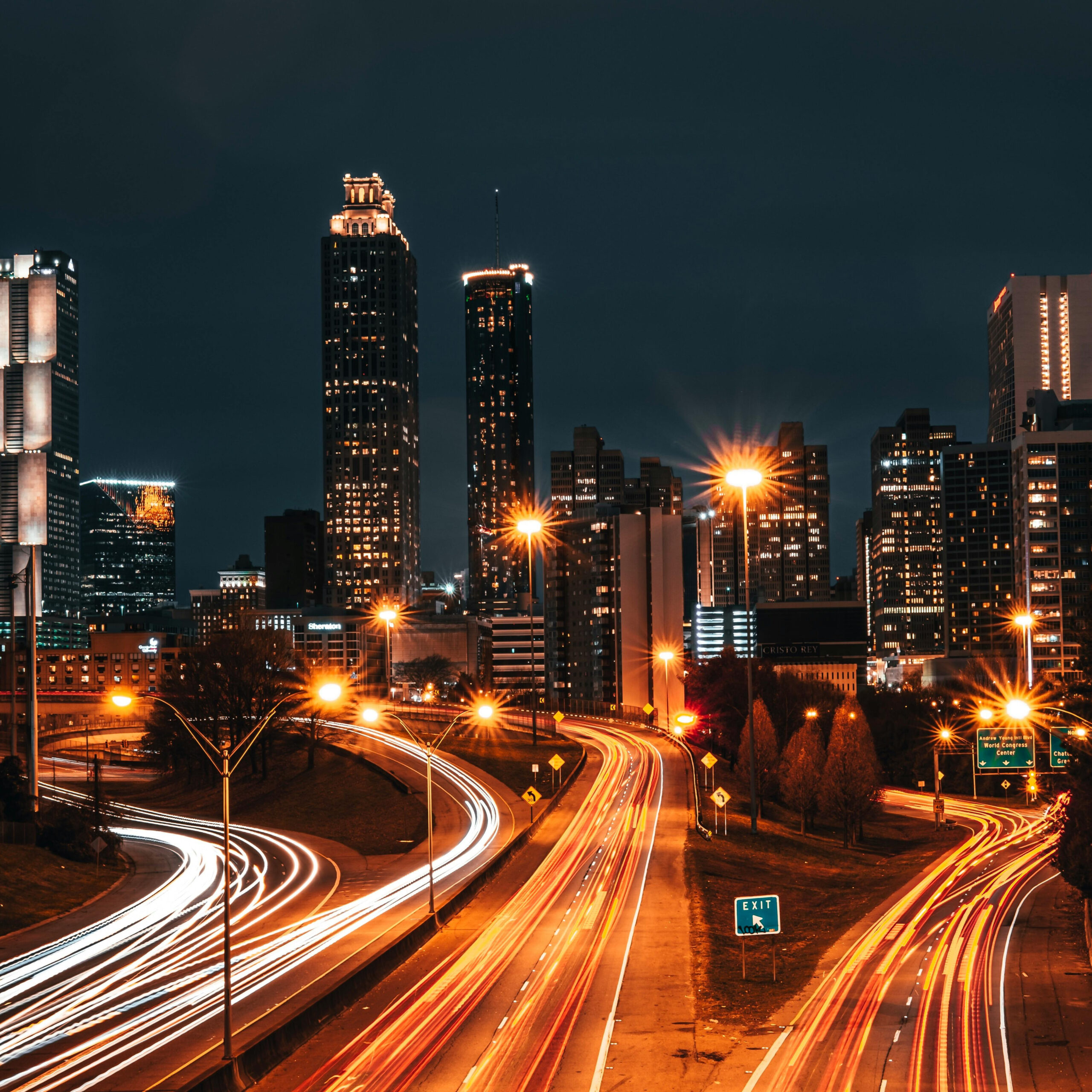 Cidade fotografada à noite com longa exposição, o trânsito formando riscos luminosos por toda a extensão da rua.